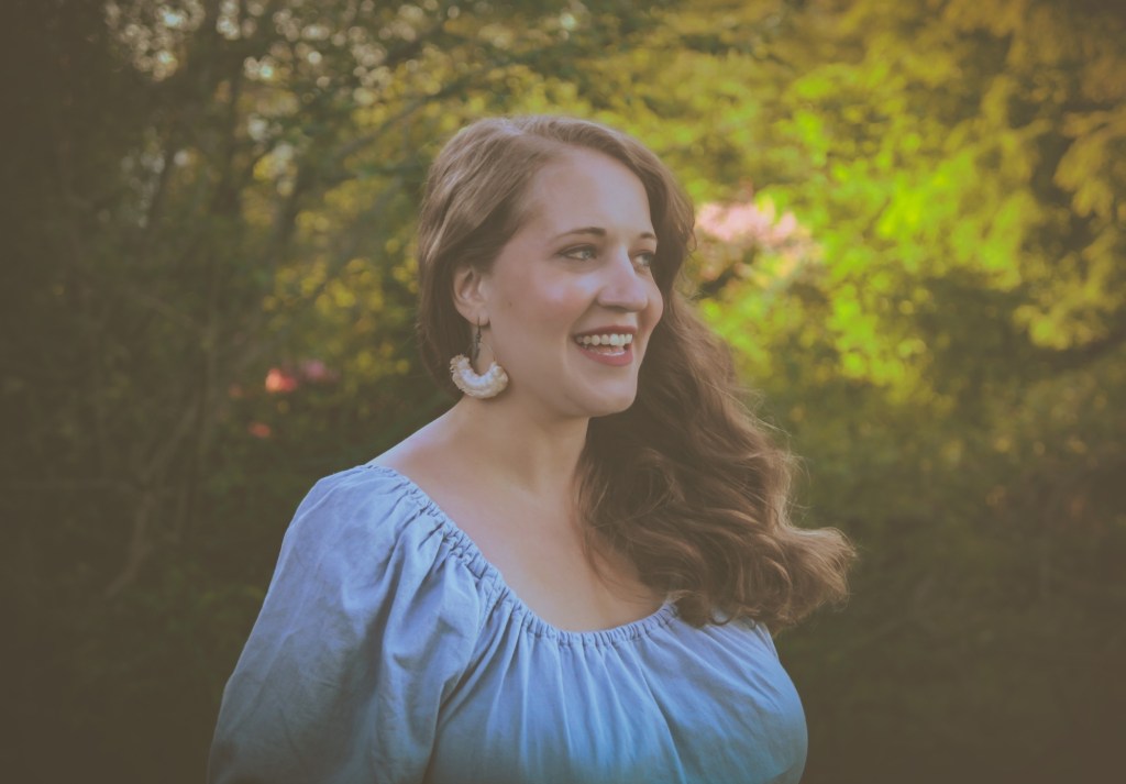 Photo by Courtney Wilkins, Jana is in a blue dress, with vibrant green trees with pink blossoms in the background. Jana has a smile on her face.