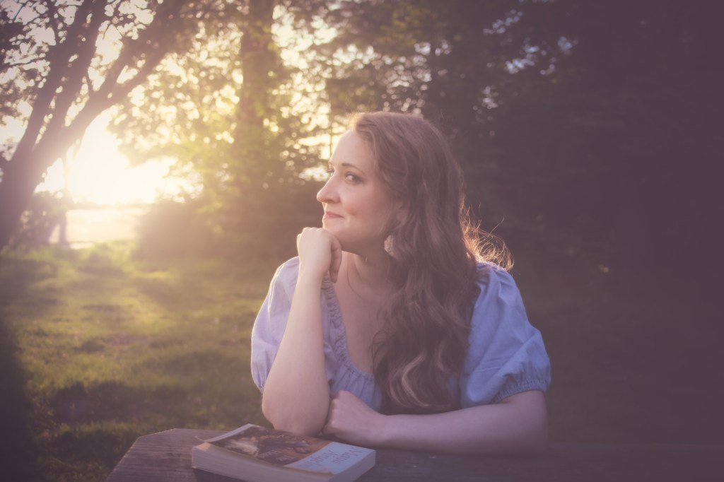 This is a photo of Jana in a blue dress, at a picnic table, with chin rested on fist pensively, and filtered sun through trees in the background.