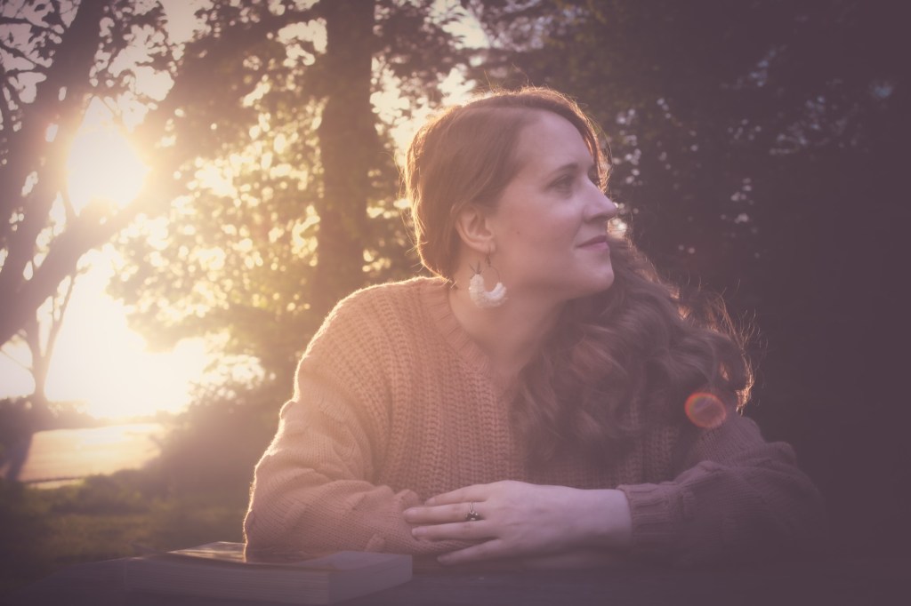 Photo by Courtney Wilkins, Jana is outside at a picnic table, wearing a blush sweater with her hair down, with sun filtering through trees.